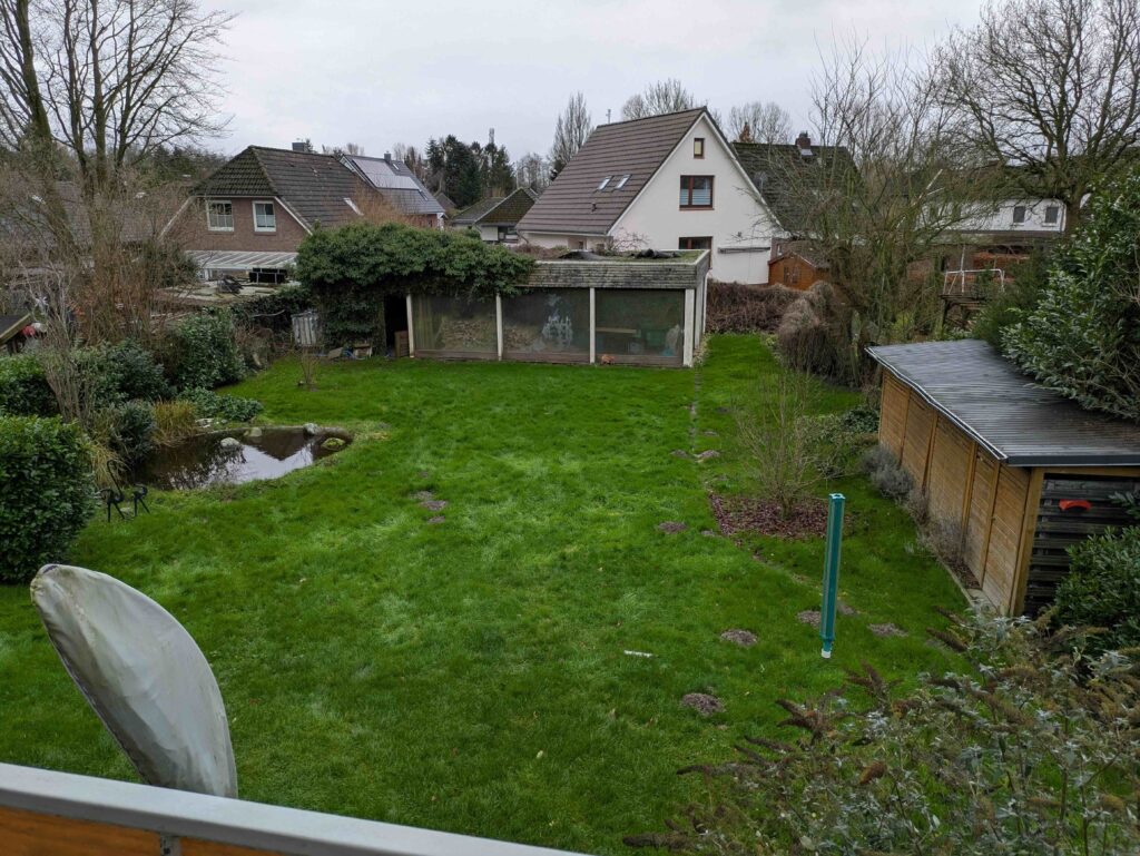 Weitwinkelaufnahme eines Gartens mit grünem Rasen, Gartenhaus und Nachbarhäusern im Hintergrund