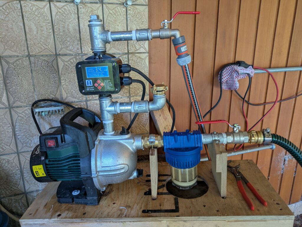 Pumpe mit Filter (blau) und Wasserdruckregler (schwarz, mit Display) und Entlüftungsöffnungen auf ein Brett geschraubt